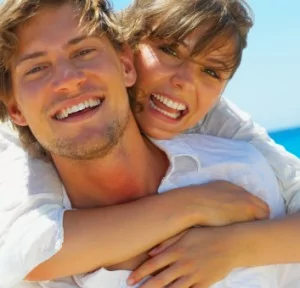 A couple holding each other on the beach, smiling confidently with white teeth.
